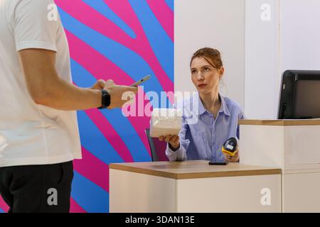 Seated woman offers small boxed item in plastic to standing man holding phone at pickup desk Stock Photo