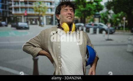 Hispanic man with headphones and hardhat stands on street clutching his back, showing discomfort while wearing casual clothing in an urban outdoor set Stock Photo
