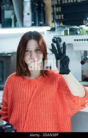 Female in glove raises three fingers in signal at modern coffee preparation area Stock Photo