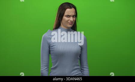 Woman with hands raised showing palms and grimace, recoil gesture, wearing blue sweater in studio against vivid green backdrop; disgust. Stock Photo