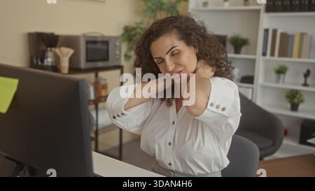 Young hispanic woman in business attire sits at a computer desk in a modern office holding her neck with both hands; work fatigue. Stock Photo