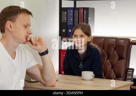 Adult male rests chin on hand thoughtfully while child smiles across office desk with cup and binders Stock Photo