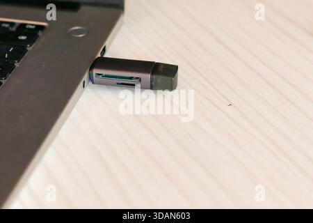 Silver laptop connected to a black multi-slot USB flash drive with SD card reader, positioned on a wooden desk for data transfer purposes. Stock Photo