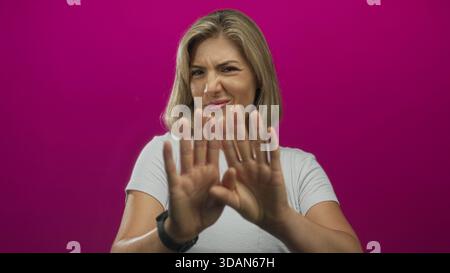 Young blonde woman scrunching nose and raising outstretched hands for stop gesture in pink studio; disgust. Stock Photo
