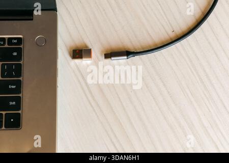A silver laptop's side view showing USB ports with a small red-accented adapter dongle and attached USB-C cable lying on a pale wooden desk. Stock Photo