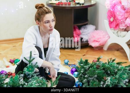 A young woman kneels on the floor, thoughtfully adjusting branches of an artificial Christmas tree amid scattered ornaments and festive decorations in Stock Photo