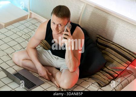 Focused young male in tank top sits on bed, intently listening to phone call with hand lightly touching silver laptop nearby Stock Photo