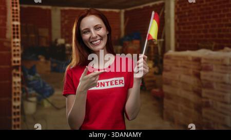 Woman smiling holding spain flag at indoor construction site highlighting volunteer work with vibrant red shirt and joyful expression in a rustic back Stock Photo