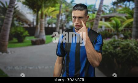 Man in blue and black soccer jersey pointing a forefinger at camera on a palm lined resort path; defiance challenge. Stock Photo
