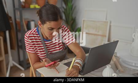 Woman holding red smartphone and writing with pen while using laptop in studio; creative concentration. Stock Photo