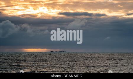 Waves gently roll in as the sun sets behind dark clouds. A hint of light shines on the water, and a distant island can be seen across the horizon, lof Stock Photo
