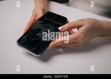 Close-up of woman removing broken tempered glass screen protector from smartphone, revealing cracked protection film on white table. Stock Photo