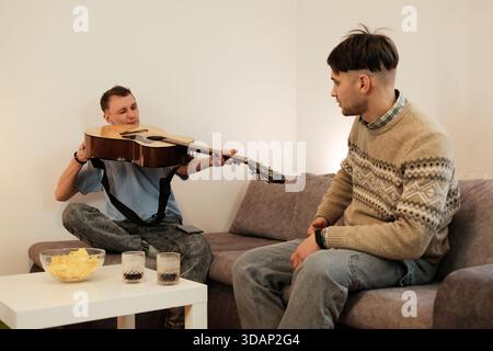 One man shows his guitar to a friend while sitting on a couch at home. The concept reflects creativity, music sharing, and friendly communication. Stock Photo