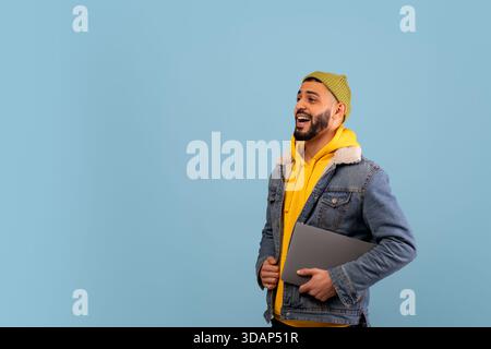 Young Man Smiles While Holding a Laptop, Dressed Casually in a Yellow Hoodie and Denim Jacket Against a Light Blue Background Stock Photo