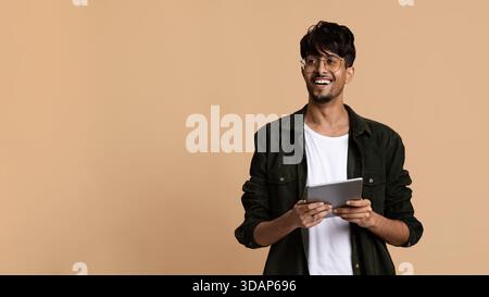Young Man Smiles While Holding a Tablet Against a Simple Beige Background in a Bright Indoor Setting Stock Photo