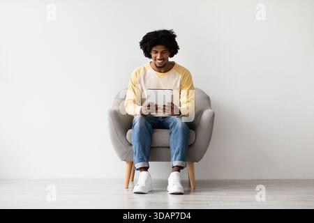 Happy Young African American Man Enjoys Digital Tablet at Home While Relaxing in Armchair Stock Photo