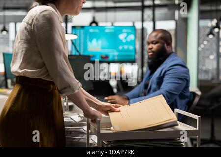 Female office assistant pushing a trolley cart with documents and folders from the archive, bringing files to a colleague and ensuring efficient distribution of resources. Administrative tasks. Stock Photo