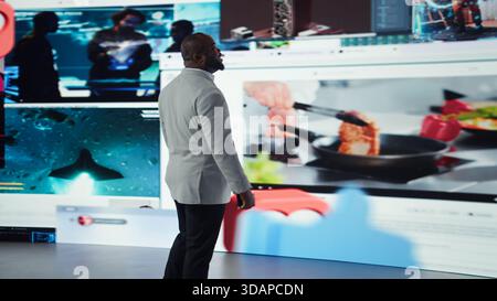 Man browsing social media feed on smartphone in front of digital video wall. Person using phone with thumbs up and heart icons floating behind him, highlighting interactive engagement online, camera A Stock Photo