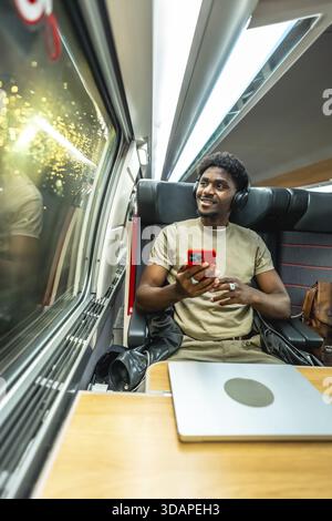Young man wearing headphones and holding a smartphone, relaxing and enjoying the journey while looking out the window of a modern train, with a laptop Stock Photo