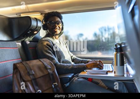 Young black man smiling while traveling on a train, wearing headphones and typing on his laptop, working remotely during a business trip or daily comm Stock Photo