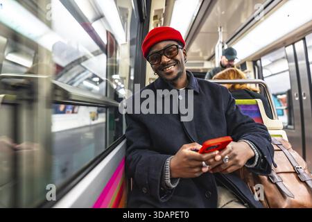 Young man smiling directly at the camera while holding and using a red smartphone, sitting on a subway train during his daily commute and journey thro Stock Photo