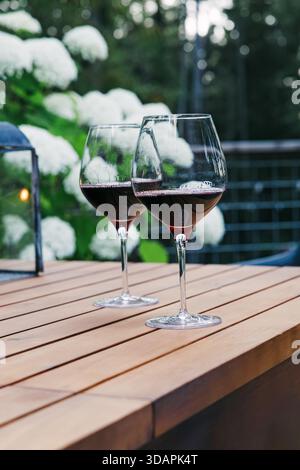 Two glasses of red wine sit elegantly on a wooden table, set against a blurred background of lush greenery and white flowers Stock Photo