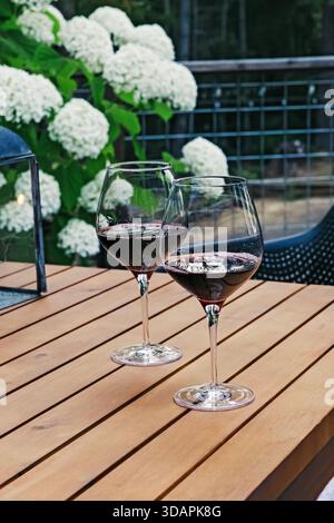 Two glasses of red wine rest on a wooden table outdoors, surrounded by lush greenery and white flowers Stock Photo
