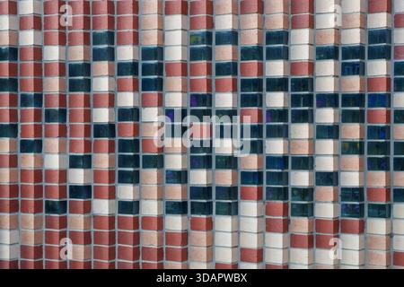 An architectural wall displays a repeating checkerboard pattern composed of textured reddish-brown bricks, light beige bricks, and translucent blue-gr Stock Photo