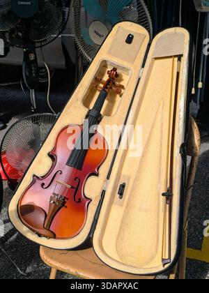 IPOH, MALAYSIA – NOVEMBER 09, 2025: A used violin and bow are displayed in an open, plush-lined case on a chair at a market stall. Stock Photo