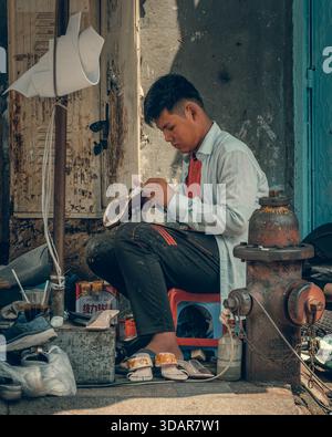 18.10.25 Ho Chi Minh Man sitting on street stool fixing a shoe in a busy Vietnamese urban environment, surrounded by tools and merchandise. Stock Photo