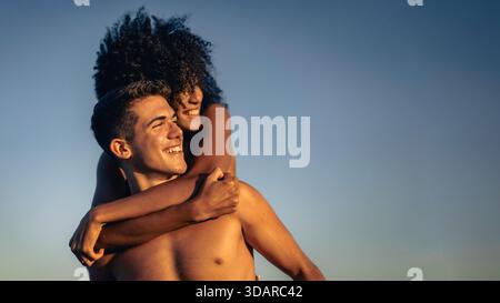 A happy interracial couple enjoying a playful piggyback ride outdoors during golden hour, their faces lit with radiant smiles against a clear blue sky Stock Photo