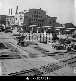 Slaviansk, Ukrainian SSR, 1970s. An archival high-angle view of the busy logistics yard at the Armature-Insulator Plant (AIZ). An industrial tractor drives across the concrete foreground, navigating between railway tracks and stacks of ceramic products. A flatbed rail wagon sits in the center, while trucks are parked at the loading docks of a massive multi-story workshop building in the background. This scene highlights the complex supply chain and economic activity of the peaceful Donbass region. Stock Photo