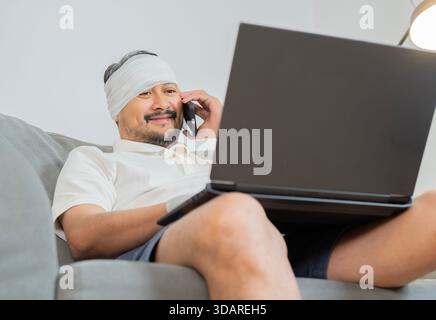 Bearded man with head injury sitting on sofa, uses laptop and phone at home for working online. Health care concept. Stock Photo