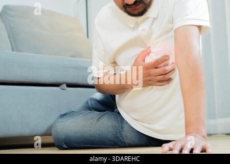 Cropped shot of Asian adult man sit on floor and holding his arms around his chest suffering from a heart attack. Heart problems concept. Stock Photo