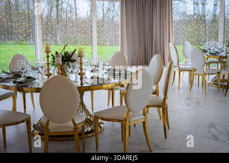 A luxurious banquet hall with round tables set for an elegant event, featuring white chairs, gold accents, and large windows Stock Photo