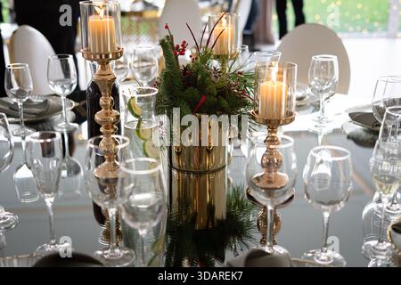 A festive dining table decorated with candles, glassware, and a centerpiece of greenery and berries in a reflective setting. Stock Photo