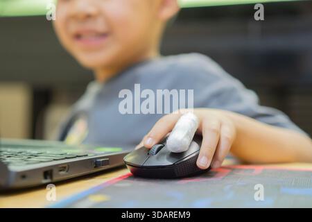 Smiling boy with bandaged on finger holding computer mouse and using laptop for play game or watching cartoon. Selective focus. Kids healthy concept. Stock Photo