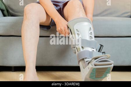 A man sitting on a soft sofa, makes adjustments and puts on an orthosis on his injured leg, demonstrating the process of home rehabilitation. Stock Photo