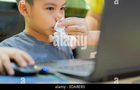Mother comforts sick child with cold, helping son to blow his nose while he using or playing on laptop at home. Kids health care. Stock Photo