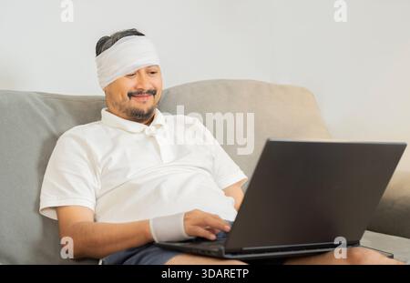 Handsome cheerful man with a broken wrist and head in bandage sitting and working at home. Trauma and rehabilitation. Social security and health insur Stock Photo
