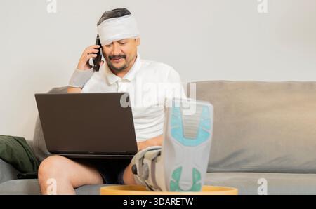 Positive man enjoying sick leave despite physical injury. Happy smiling young guy with broken leg and head sitting in sofa at home, using mobile phone Stock Photo