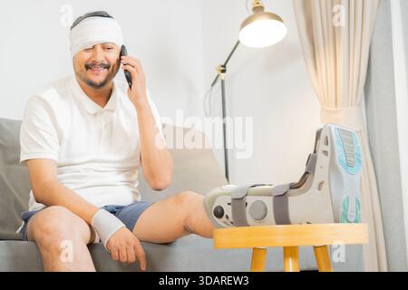 Sick adult Asain man sit on sofa wearing bandages his head, wrist and leg using smartphone at home. Health care concept. Stock Photo