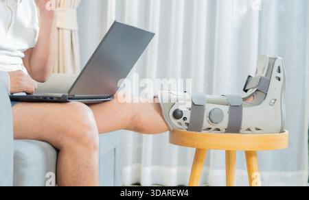 Side view shot of man with broken leg working on laptop at home sitting on sofa with crutches after accident. Male injured person using computer on re Stock Photo