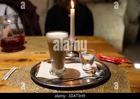 Glass of latte and water served on a silver tray beside a lit candle, creating a warm and cozy atmosphere on a rustic wooden table. Stock Photo