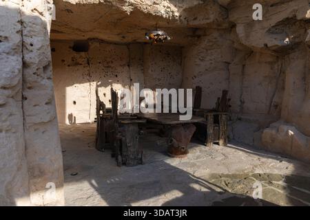 Cozy stone-walled cave interior with wooden dining table and chairs bathed in natural light, offering a unique and atmospheric gathering space. Stock Photo