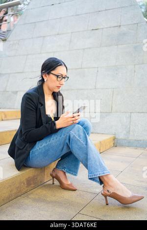Woman sitting on concrete stairs, focused on her smartphone Stock Photo
