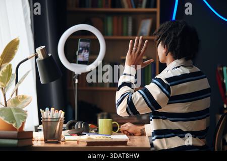 Young adult Black man sitting at desk waving hand while recording video or participating in online class on smartphone mounted on ring light, surrounded by books and stationery at home Stock Photo