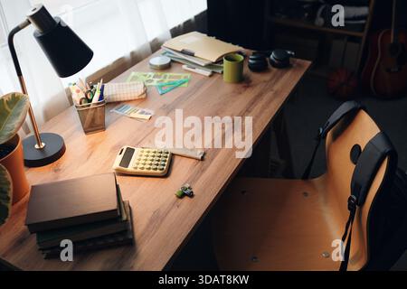 Wooden desk holding calculator, books, stationery, green mug, headphones, and plant near window, empty chair with backpack hanging on backrest, lamp light illuminating workspace Stock Photo