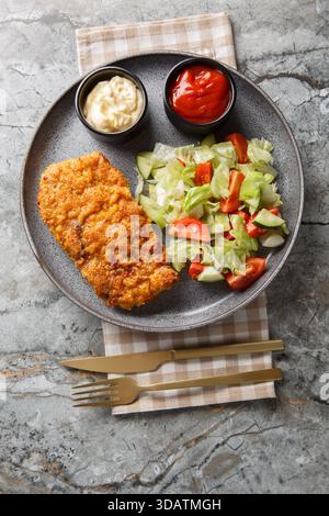 Sicilian breaded beef steak served with fresh salad and two sauces close-up on a plate on the table. Vertical top view from above Stock Photo