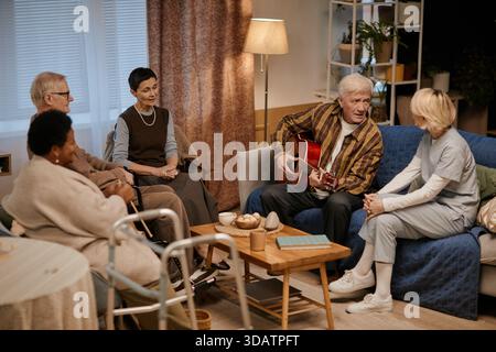 Group of Senior Residents of Nursing Home Listening to Man Playing Guitar Stock Photo